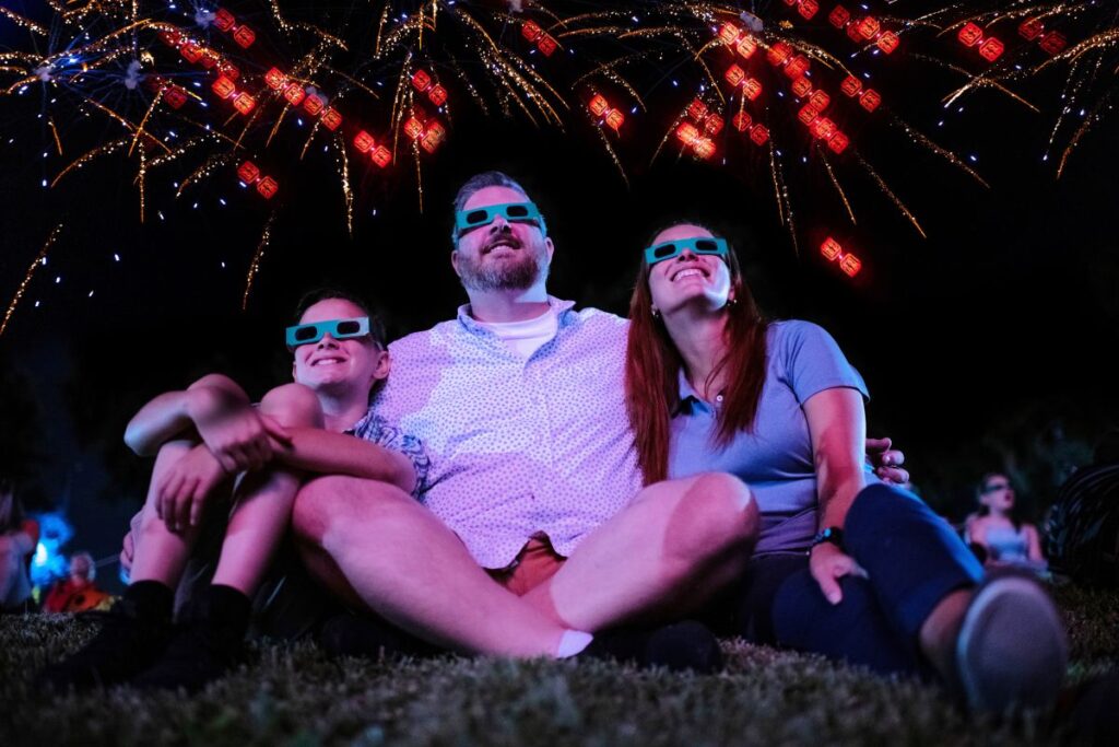 A family of three—a boy, man, and woman—sit on the grass at night, looking up and smiling while wearing 3D glasses. Red, glowing fireworks shaped like Lego bricks burst across the dark sky above them for Red, White & BOOM!