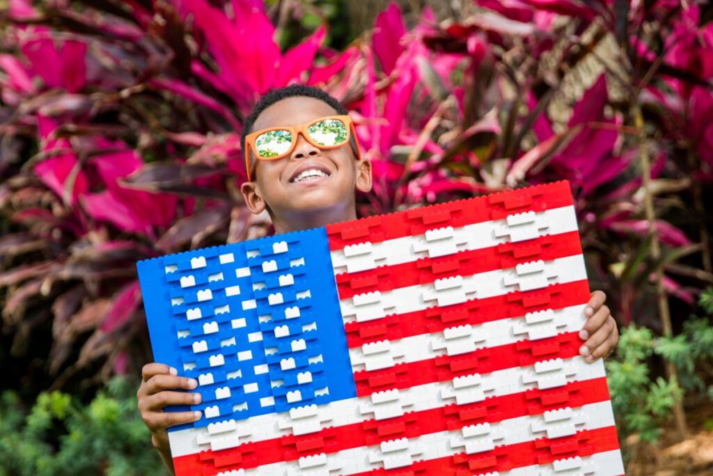 A young boy wearing bright orange sunglasses smiles up toward the sky while holding a large American flag built entirely out of red, white, and blue Lego bricks. Vibrant pink and red foliage fills the background.