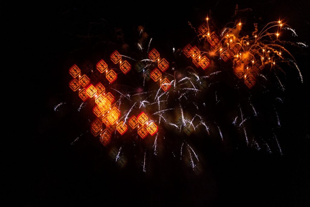 Fourth of July Legoland - A close-up view of fireworks bursting in a dark night sky, transformed into glowing, three-dimensional red and orange Lego brick shapes through a special lens or 3D glasses.