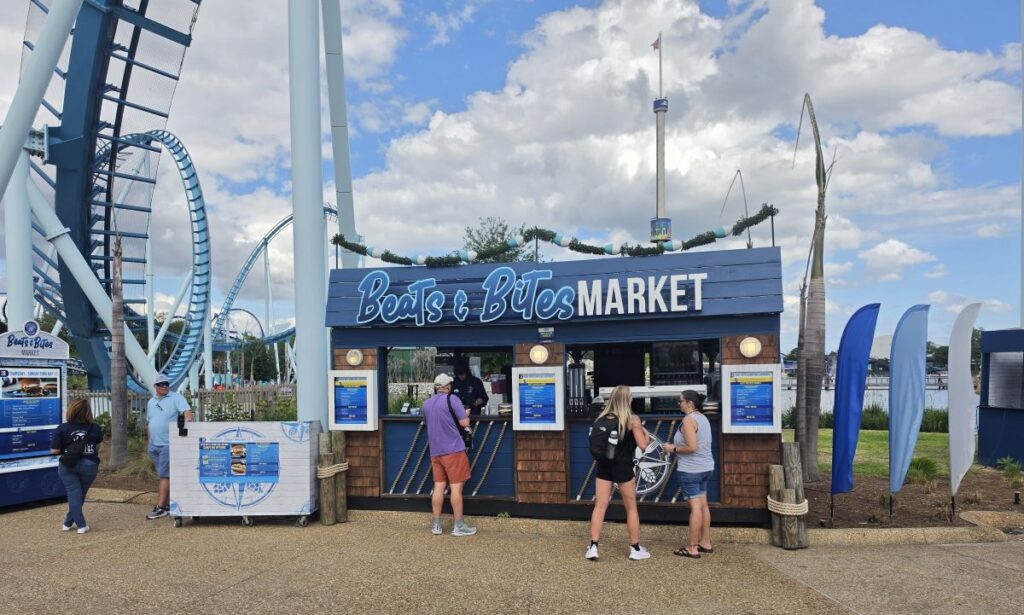Beats & Bites Market kiosk at an amusement park with a roller coaster in the background and people ordering food on a sunny day.