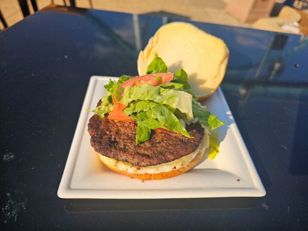 Hamburger with lettuce, tomato, and onion on a white plate with the top bun nearby on a dark table.