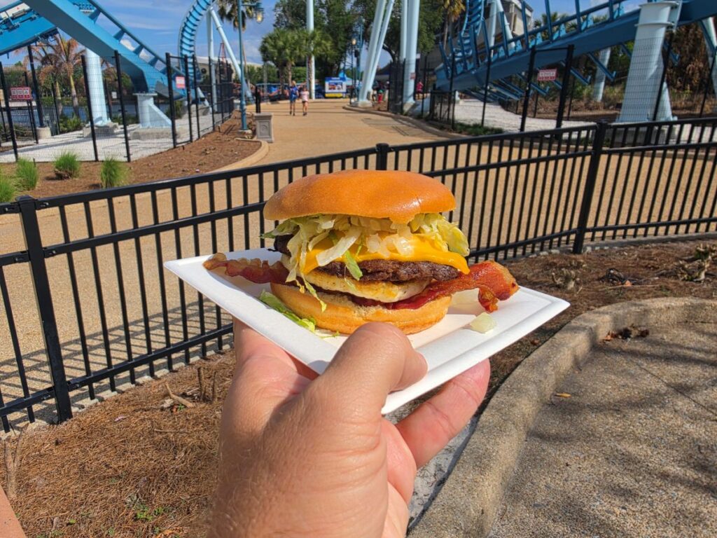 Hand holds bacon cheeseburger on a white tray in front of a blue roller-coaster at an amusement park for Seven Seas Food festival