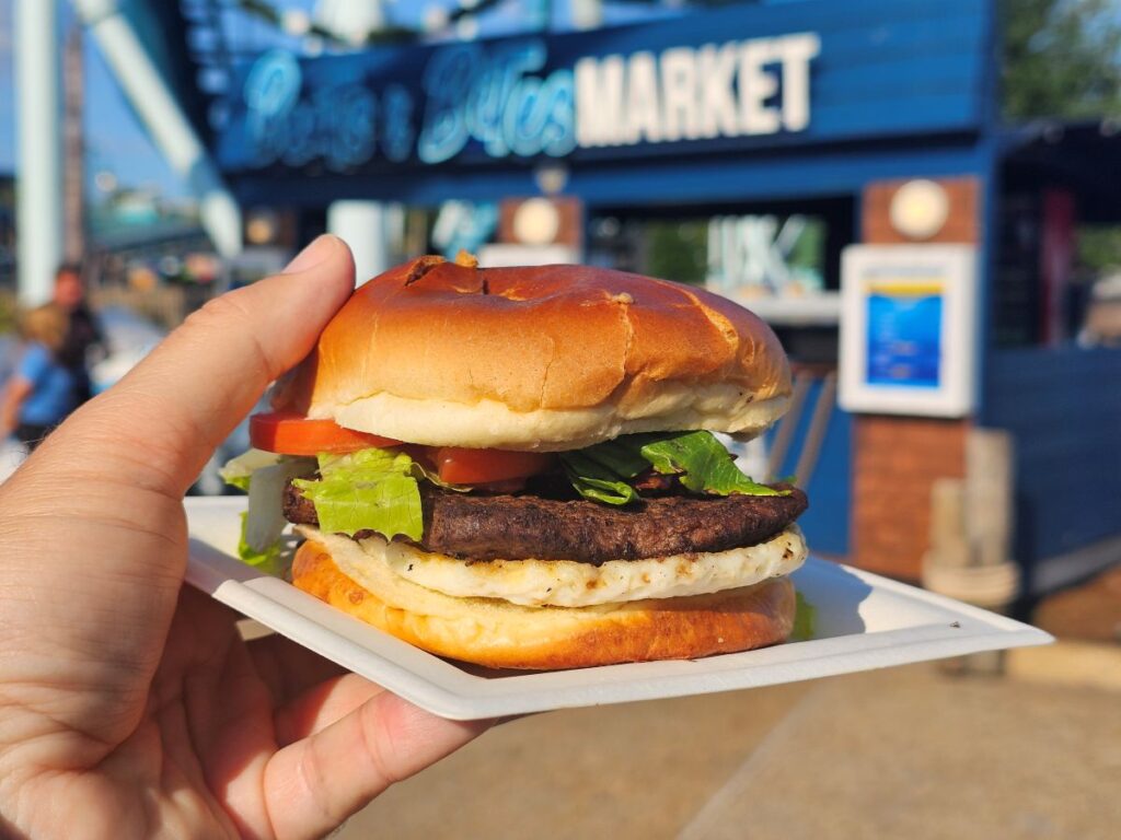 Hand holding a hamburger on a white plate, with lettuce, tomato, and beef patty, in front of a blue market building.