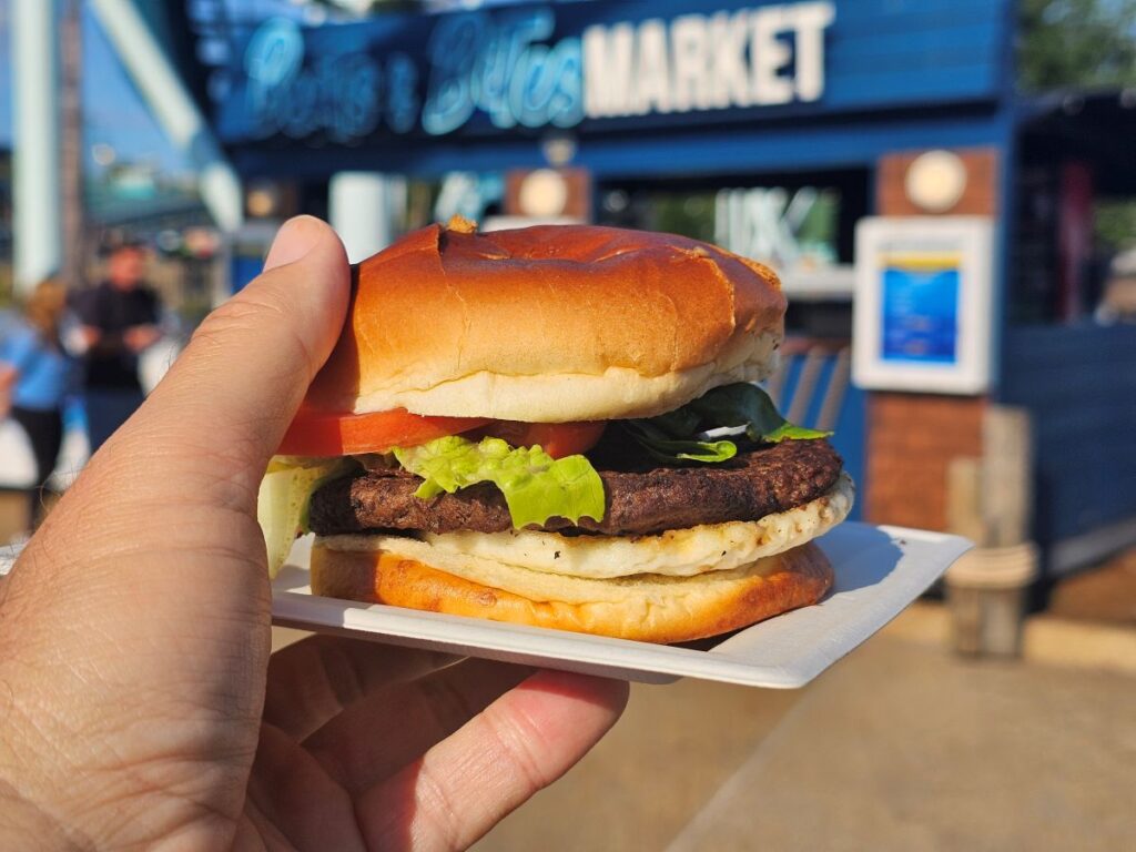Hand holding a beef burger with lettuce and tomato at an outdoor market.