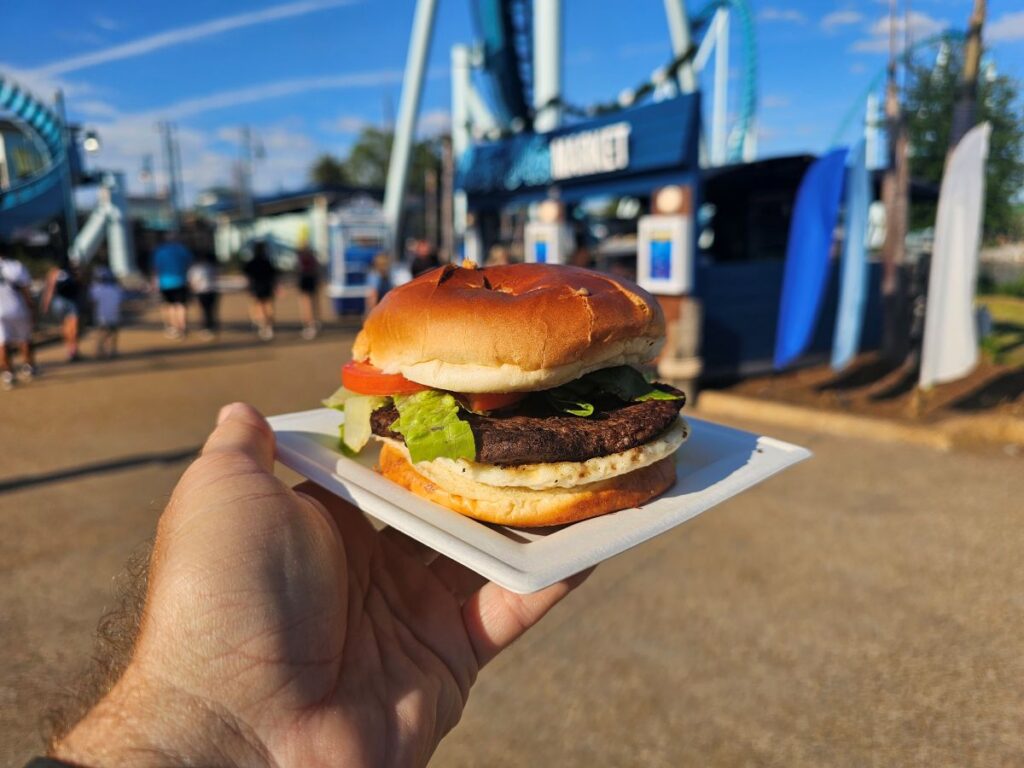 Breakfast Burger Review: Close-up of a beef burger with lettuce and tomato on a white plate, held up in front of a blue-sky amusement park with roller coasters in the background.