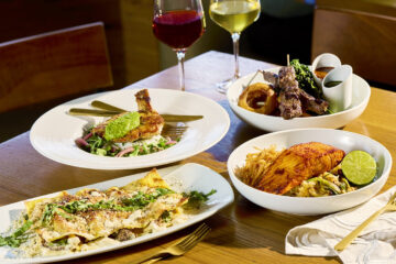 Table of food on a wooden table: pasta with greens, roasted chicken, salmon dish with lime, onion rings, salad, and two wine glasses (red and white).