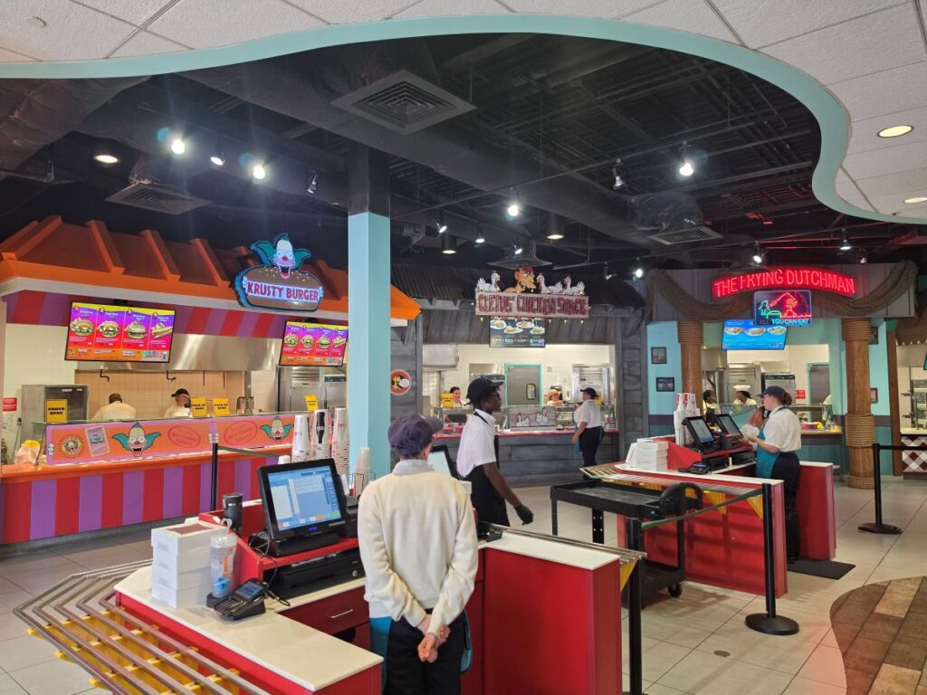 Bright fast-food restaurant interior with a Krusty Burger sign, order counters, and staff taking orders.