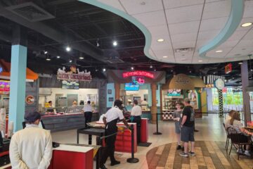 Food court scene with counter-service stalls, including signs for The Frying Dutchman and Luigi's, as customers order and dine nearby.