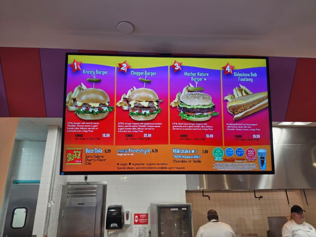 Bright Fast Food Boulevard menu board displaying four burgers: Krusty Burger, Clogger Burger, Mother Nature Burger, Sideshow Bob Footlong, with fries on the side.