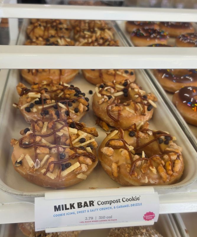 Six glazed donuts topped with chocolate drizzle, nuts, and cookie pieces in a bakery case labeled 'MILK BAR Compost Cookie'