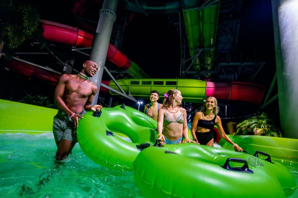 Four friends in swimwear push a bright green inflatable raft through a pool at a water park, with colorful slides in the background in neon lights.