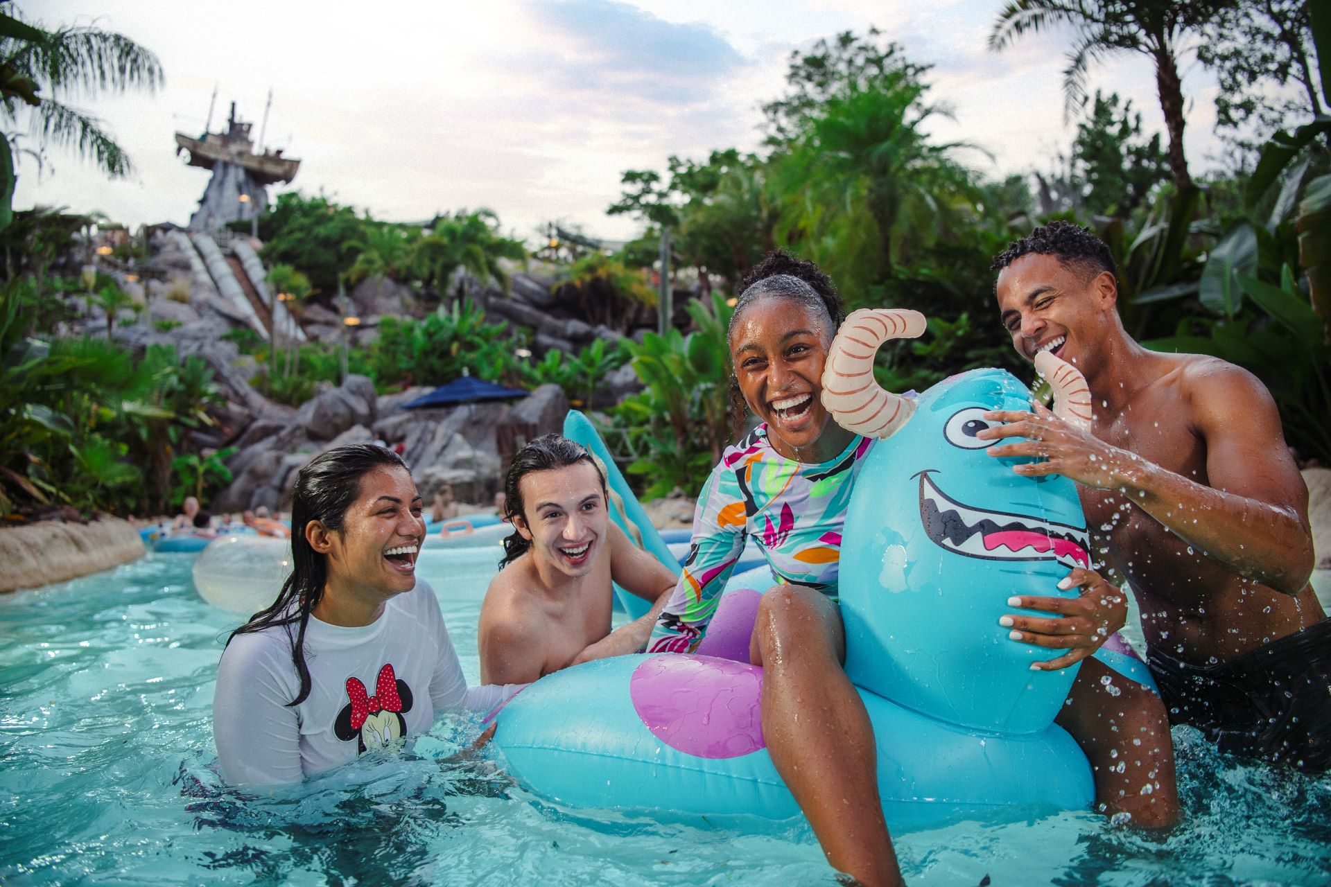 Group of friends laughing on a blue inflatable raft in a tropical water park setting with green plants and rocks in the background. News of Typhoon Lagoon Closing Date