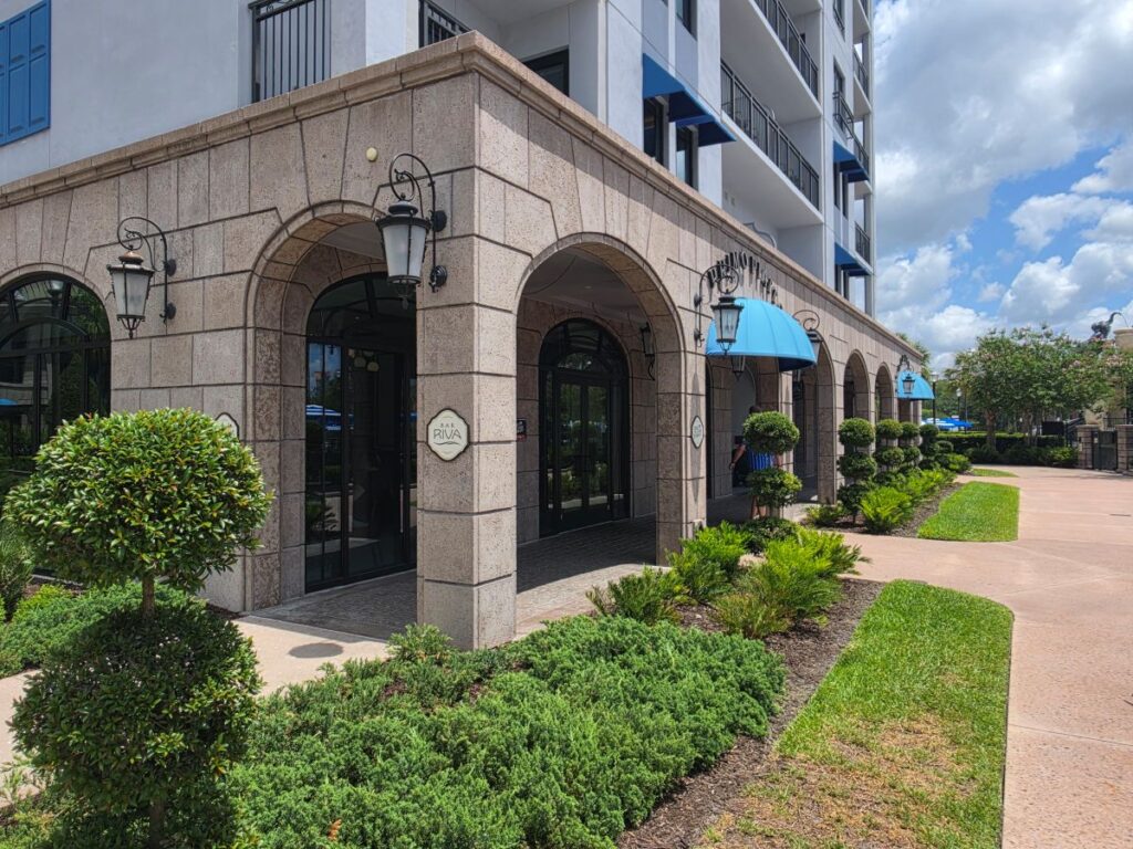 Hotel entrance with stone arches, glass doors, and blue fabric canopies; landscaped walkway with shrubs nearby.