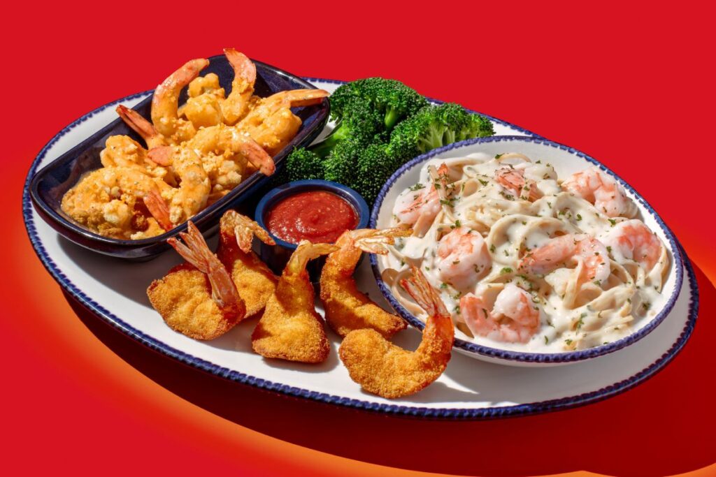 Seafood platter with fried shrimp, shrimp Alfredo pasta, broccoli, and a dipping sauce on a white platter against a red background.
