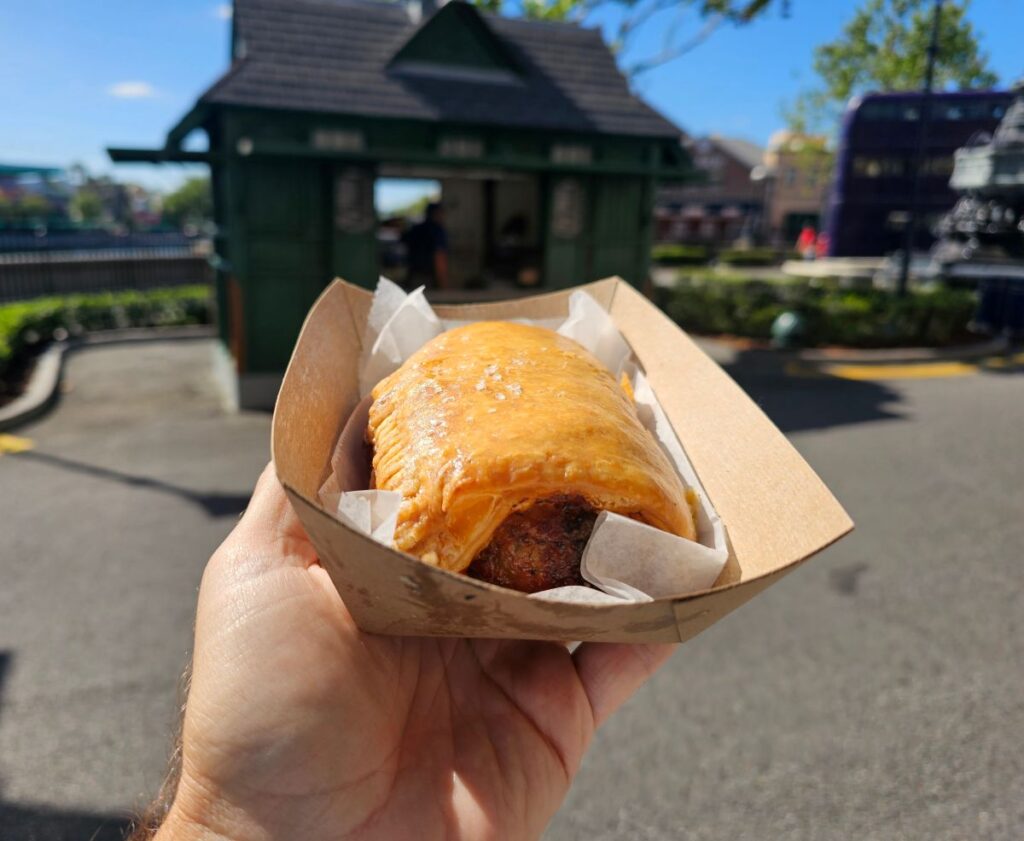 Hand holding a flaky sausage roll in a takeout box outdoors on a sunny day.