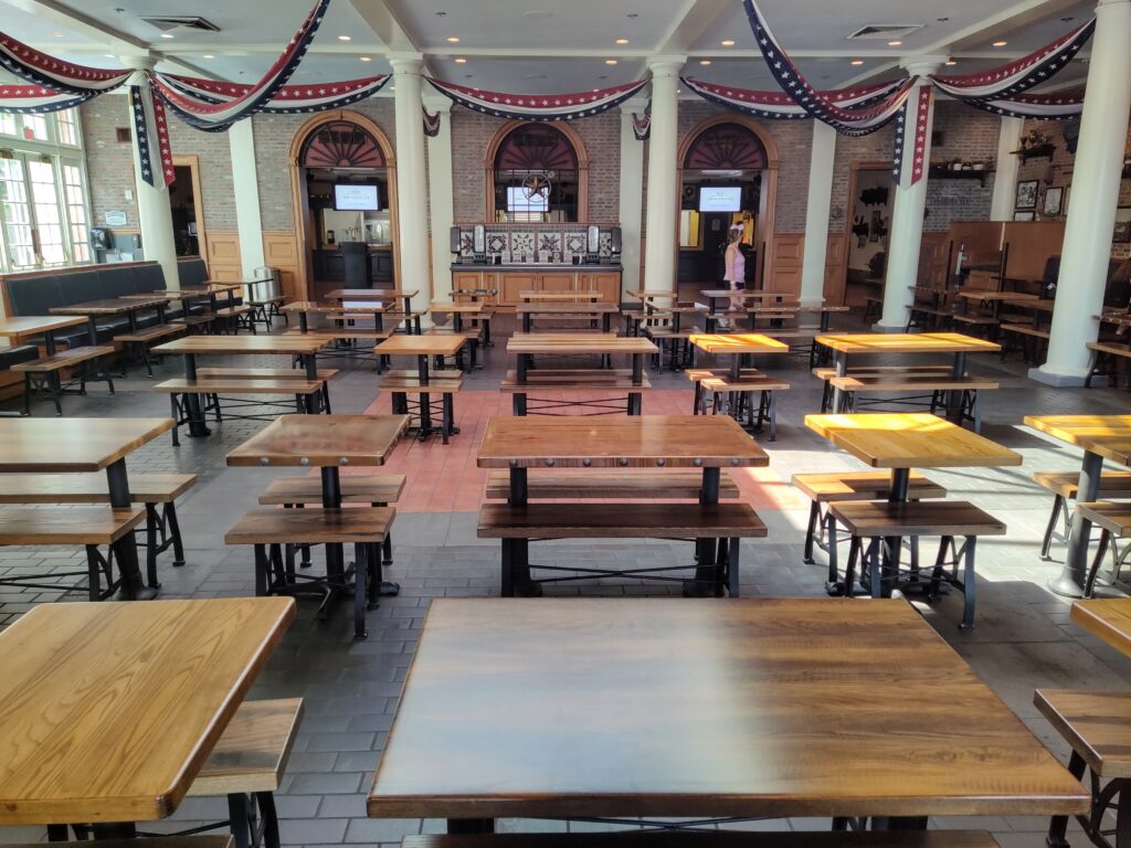 Large hall set up as a communal dining area with many wooden tables and benches, red-white-blue bunting overhead, brick walls, and a person walking near the back.