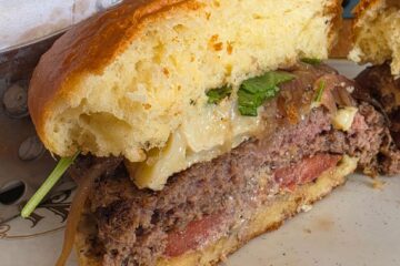 Close-up of a juicy cheeseburger cut in half on a plate, showing a beef patty, melted cheese, onions, greens, and a toasted bun with fries nearby