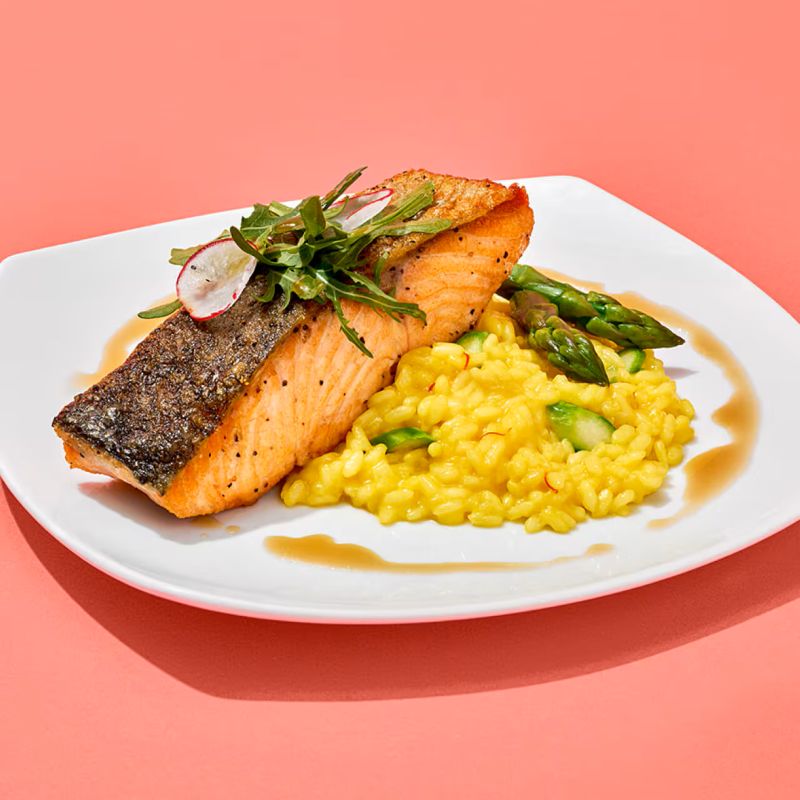 Seared salmon fillet placed over creamy saffron risotto with asparagus, garnished with greens on a white plate.