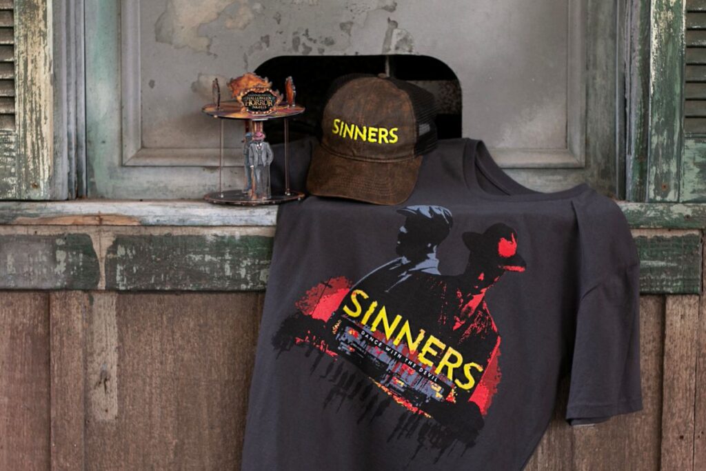 Gray T-shirt with a bold yellow 'SINNERS' graphic, draped over a rustic windowsill; a brown cap also labeled 'SINNERS' sits nearby with a small horror-themed figurine display beside it.