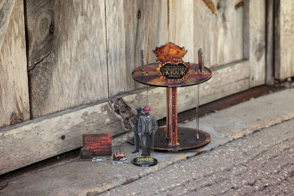 Outdoor Halloween display on a sidewalk: a round rusty metal table with a Halloween Horror Nights sign, plus small figurines and props near a weathered wooden wall.