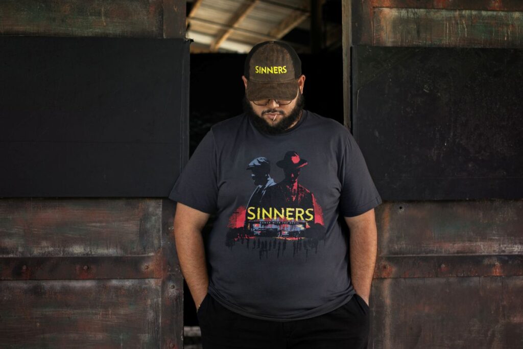 Man with a beard stands in a dark doorway, wearing a black cap with 'Sinners' and a graphic t‑shirt that also reads 'Sinners', hands in pockets.