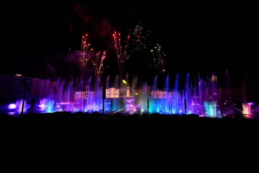 Night scene of a colorful fountain show with rainbow-lit water jets and fireworks above the silhouette of a stage or plaza buildings in the background