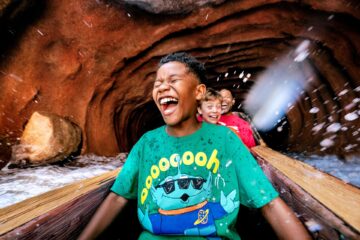 Children ride a log flume through a rocky tunnel, laughing as water splashes around the boat.