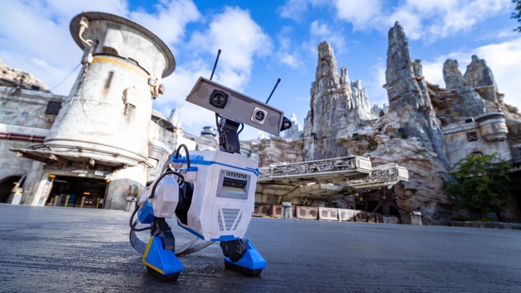 Robot with a rectangular sensor head and blue-and-white body stands on a paved surface in a rugged sci‑fi landscape under a blue sky.