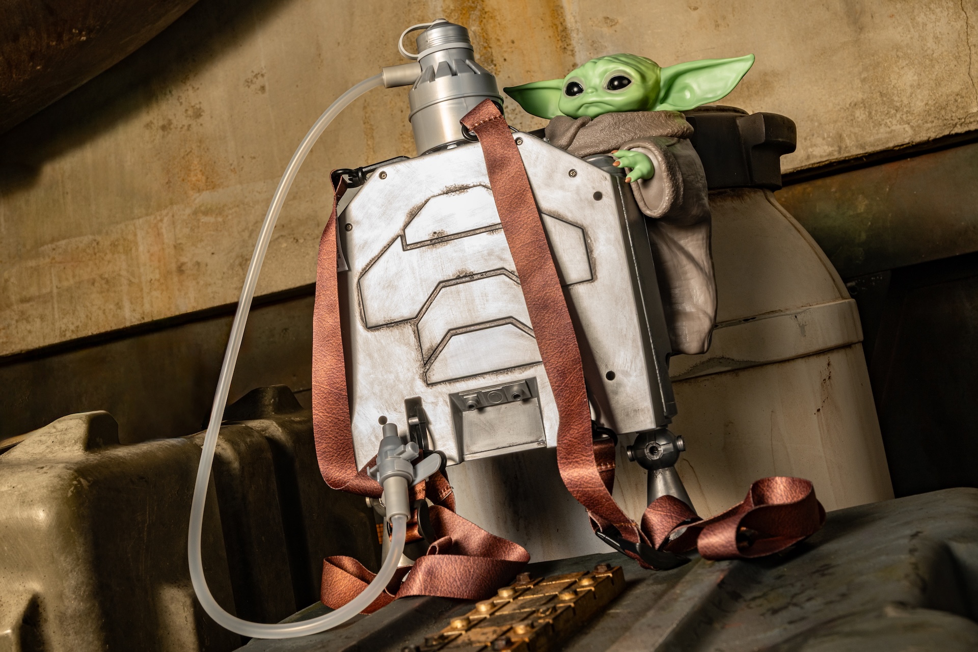 Baby Yoda (Grogu) perched on a weathered metal jetpack with brown straps in an industrial setting
