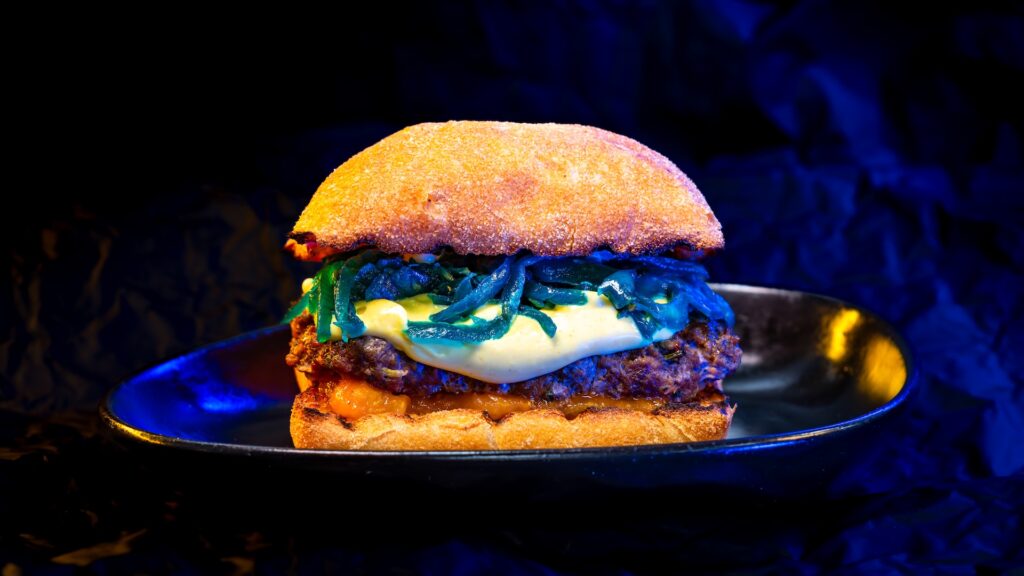 Close-up of a hamburger with melted cheese and sautéed greens on a toasted bun, on a dark plate under dramatic lighting.
