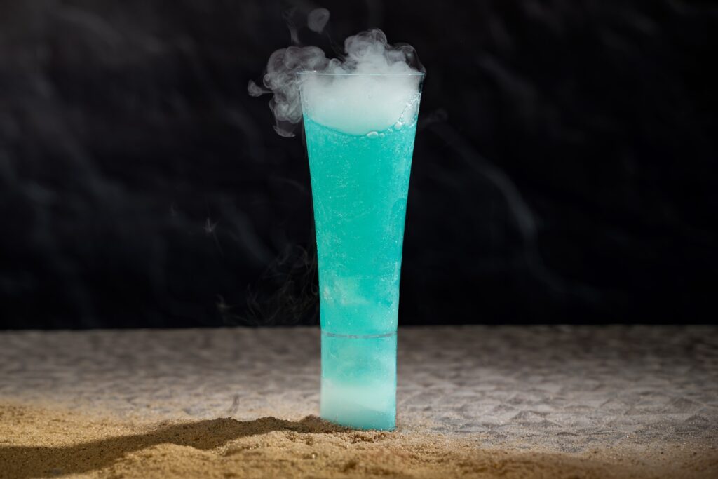Tall turquoise cocktail in a long glass with smoky fog spilling over the rim on a sandy beach at night.