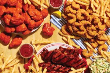 Assorted fried foods on a wooden table for Applebee's All You Can Eat promotion: crispy chicken bites, french fries, ribs with barbecue sauce, and dipping sauces with a small lemon wedge nearby.