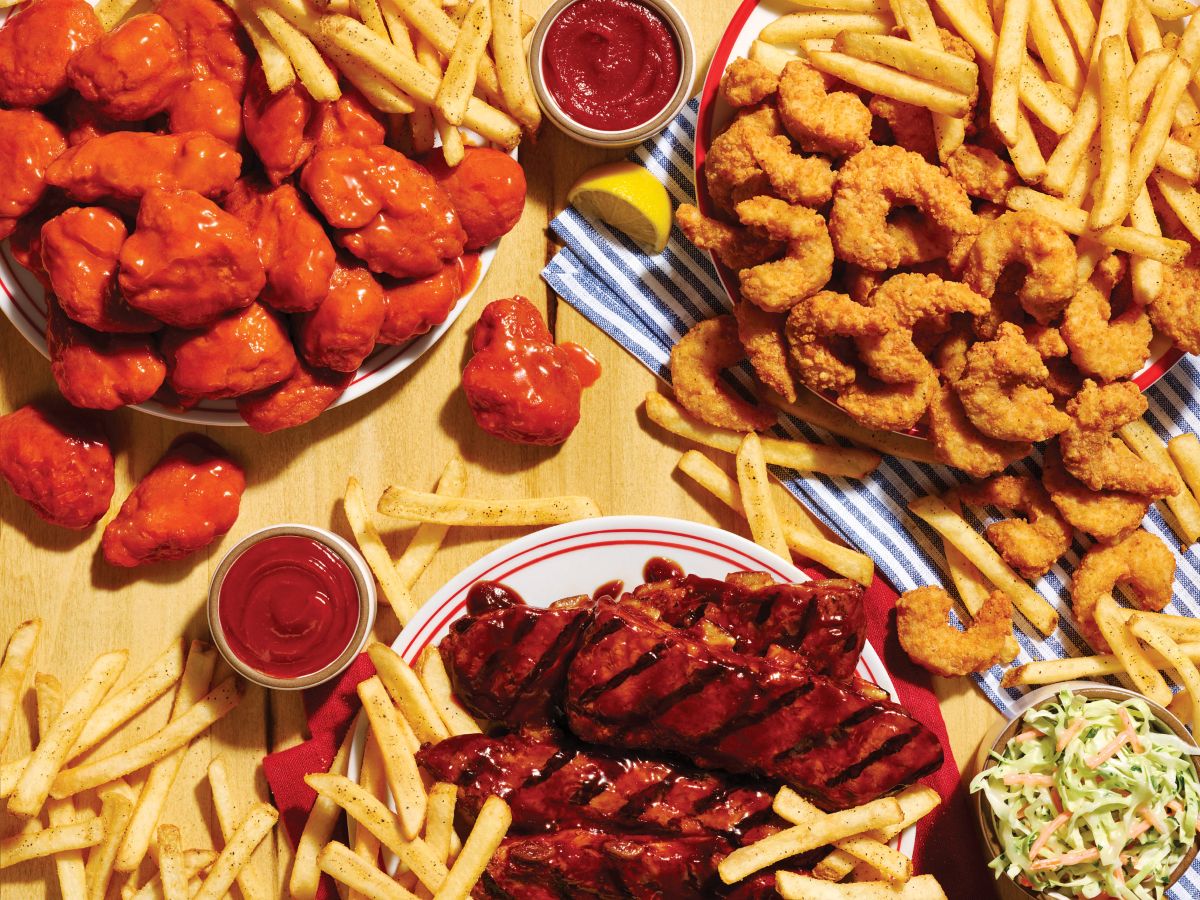 Assorted fried foods on a wooden table for Applebee's All You Can Eat promotion: crispy chicken bites, french fries, ribs with barbecue sauce, and dipping sauces with a small lemon wedge nearby.