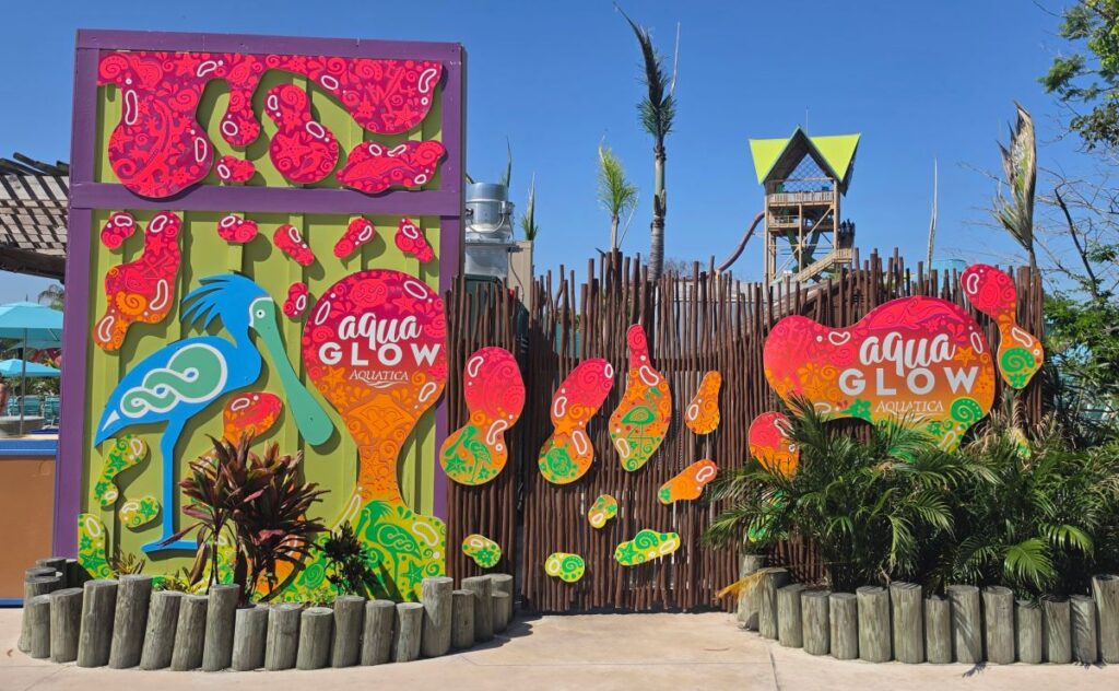 Colorful water park entrance with a bright mural reading 'aqua GLOW Aquatica' and a wooden fence backdrop, blue sky above.