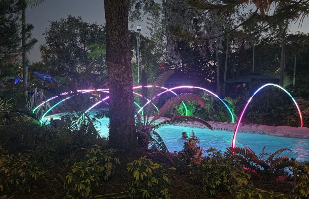 Night pool area at dusk with colorful neon arches arching over a lit pool, surrounded by tropical plants and trees.