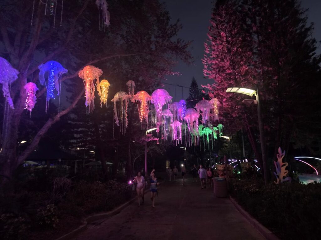 Night scene of colorful jellyfish-shaped light lanterns hanging above a walkway in a park at night, with people strolling beneath them for AquaGlow Aquatica Orlando