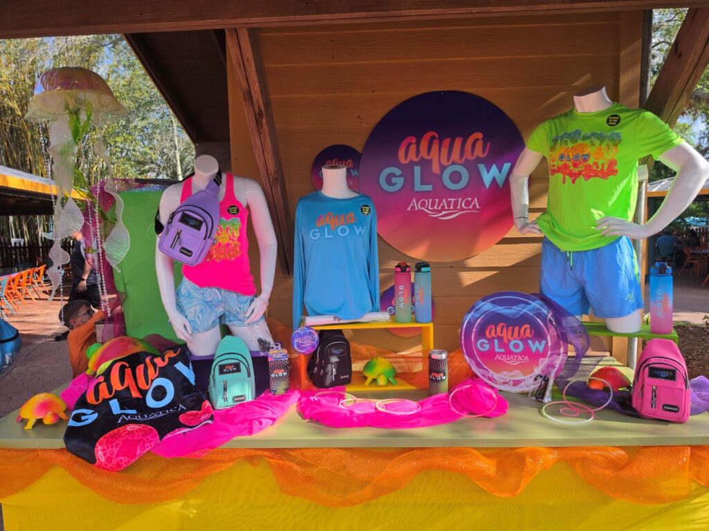 Colorful Aqua Glow Aquatica merch display with neon shirts on mannequins, backpacks, water bottles, and accessories on a table at a booth.