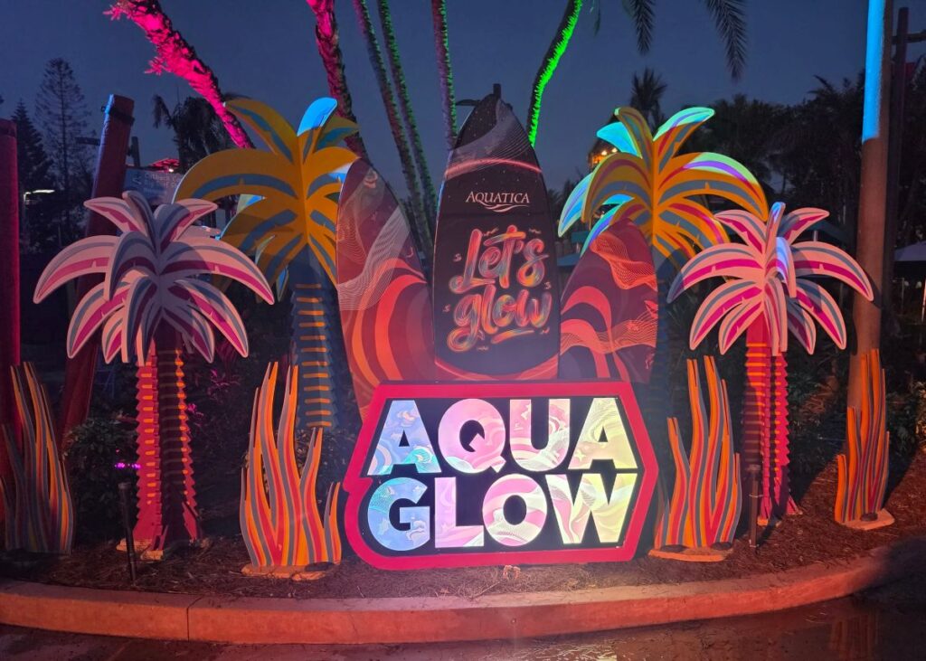 Colorful neon tropical display at night with palm trees and a glowing sign reading AQUA GLOW and Let's Glow attributed to Aquatica for AquaGlow Aquatica Orlando