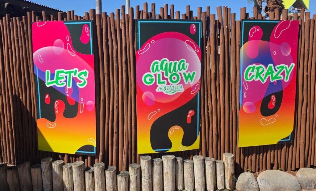 Three colorful posters on a brown wooden fence advertising Aqua Glow with bold words: Let's, Aqua Glow, Crazy.
