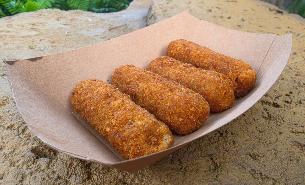Five golden-brown breaded croquettes arranged in a paper tray on a sandy surface.