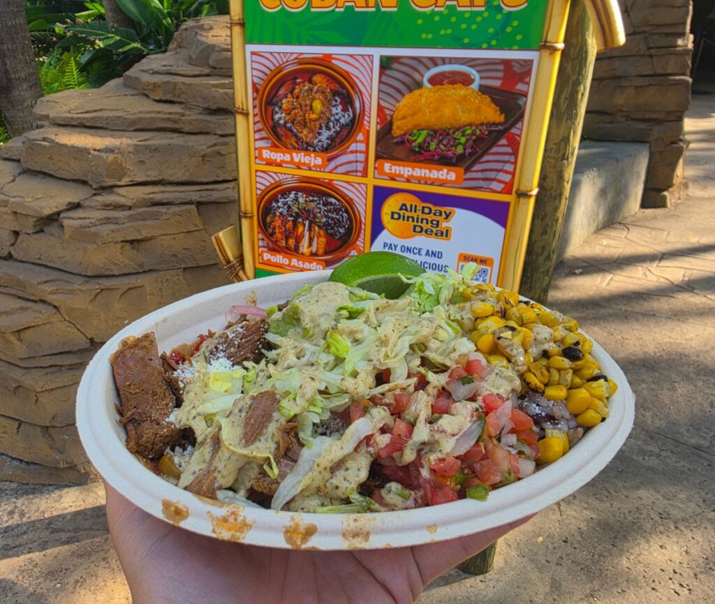 Disposable bowl piled with lettuce, tomatoes, corn, grilled meat, and creamy sauce, held outdoors beside a colorful menu sign for BG Cuban Cafe.