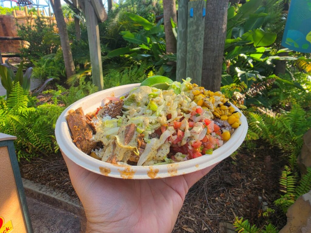 Hand holding a white foam bowl filled with a colorful veggie-topped taco/bowl, including beef, cheese, rice, lettuce, tomatoes, corn, and guacamole, outdoors among lush plants.