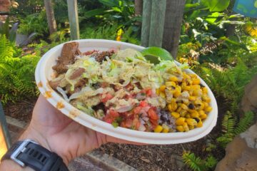 Disposable takeout bowl with mixed toppings (corn, lettuce, tomatoes, beans) and lime, held outdoors by a person wearing a black wristwatch