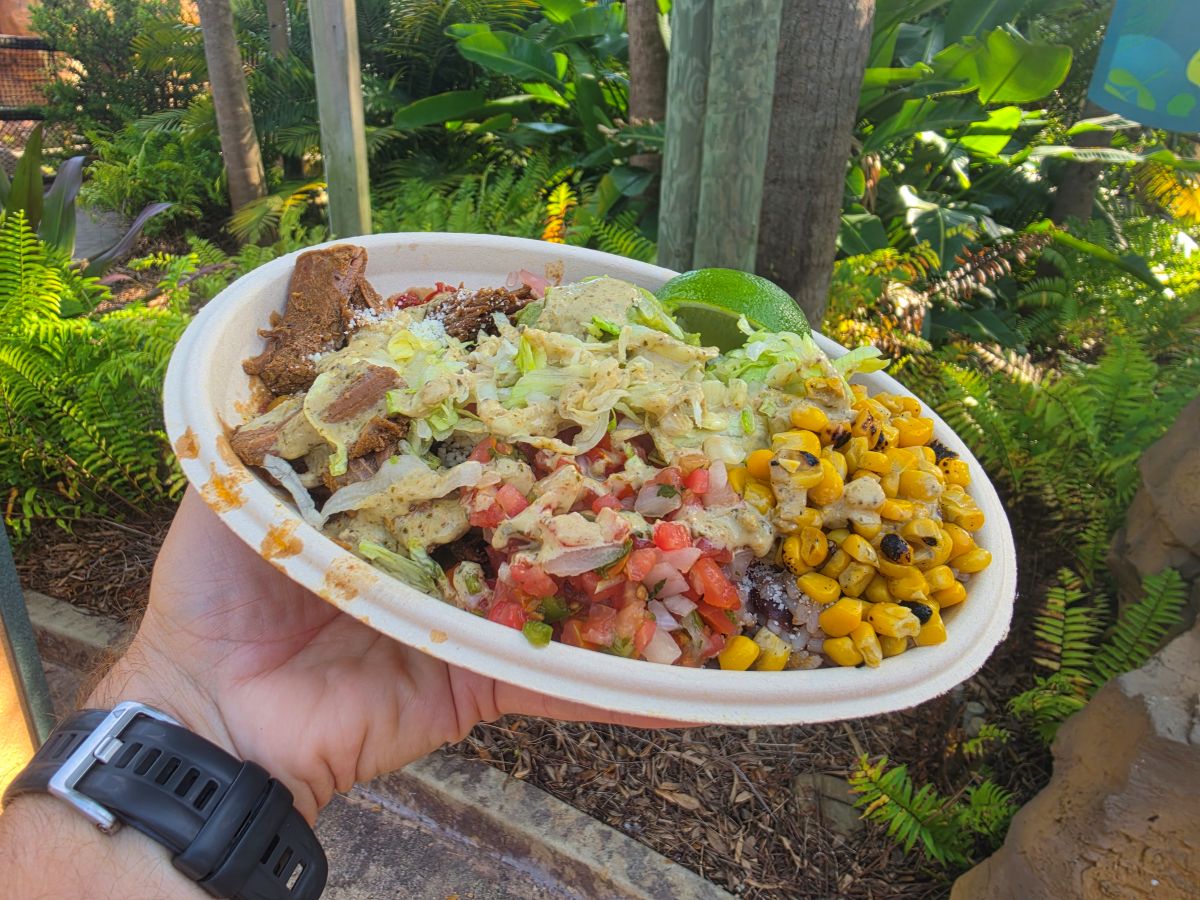 Disposable takeout bowl with mixed toppings (corn, lettuce, tomatoes, beans) and lime, held outdoors by a person wearing a black wristwatch