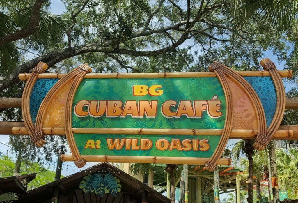 Bright sign reading 'BG Cuban Café at Wild Oasis' hanging from a wooden bamboo frame with tropical background