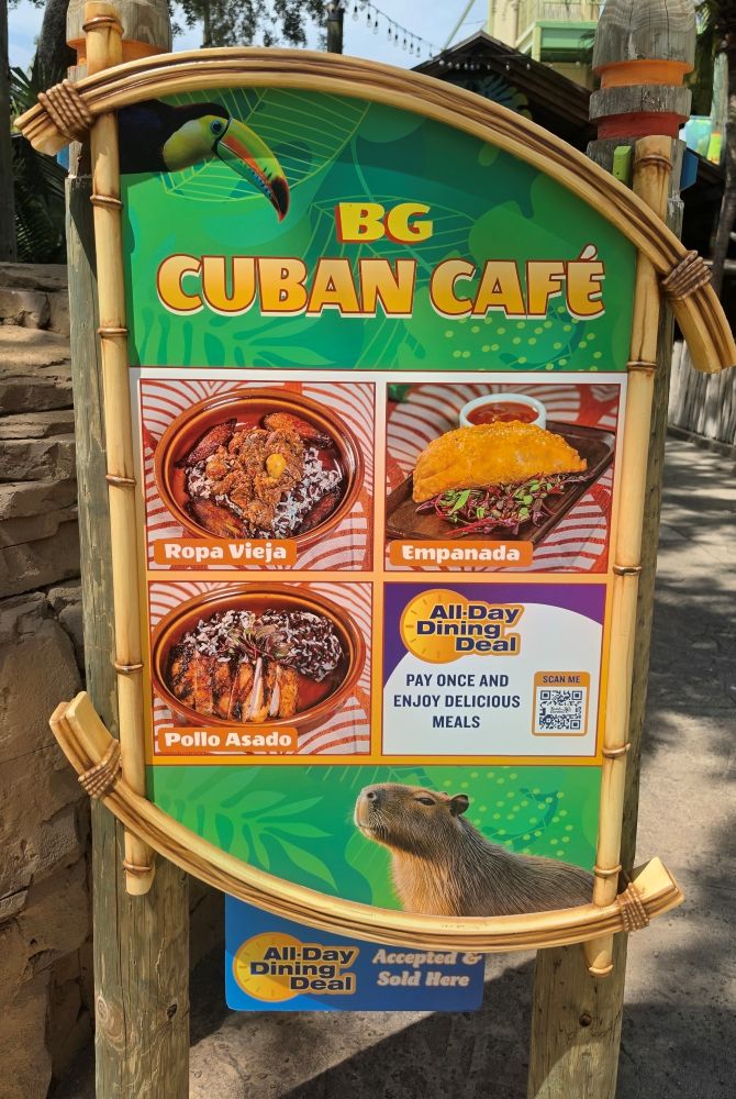 Outdoor sign for BG Cuban Café with a toucan graphic and tropical green background, displaying menu photos of Ropa Vieja, Empanada, and Pollo Asado and a capybara illustration at the bottom (All-Day Dining Deal).