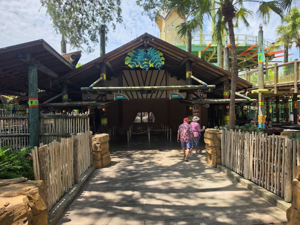 Two older adults walk toward the entrance of a rustic, tropical-themed pavilion with palm trees and colorful posts.