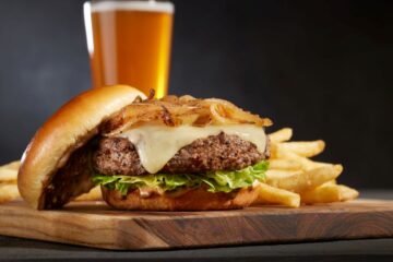 Cheeseburger with melted cheese, lettuce, and caramelized onions on a toasted bun, with fries and a beer in the background on a wooden board.