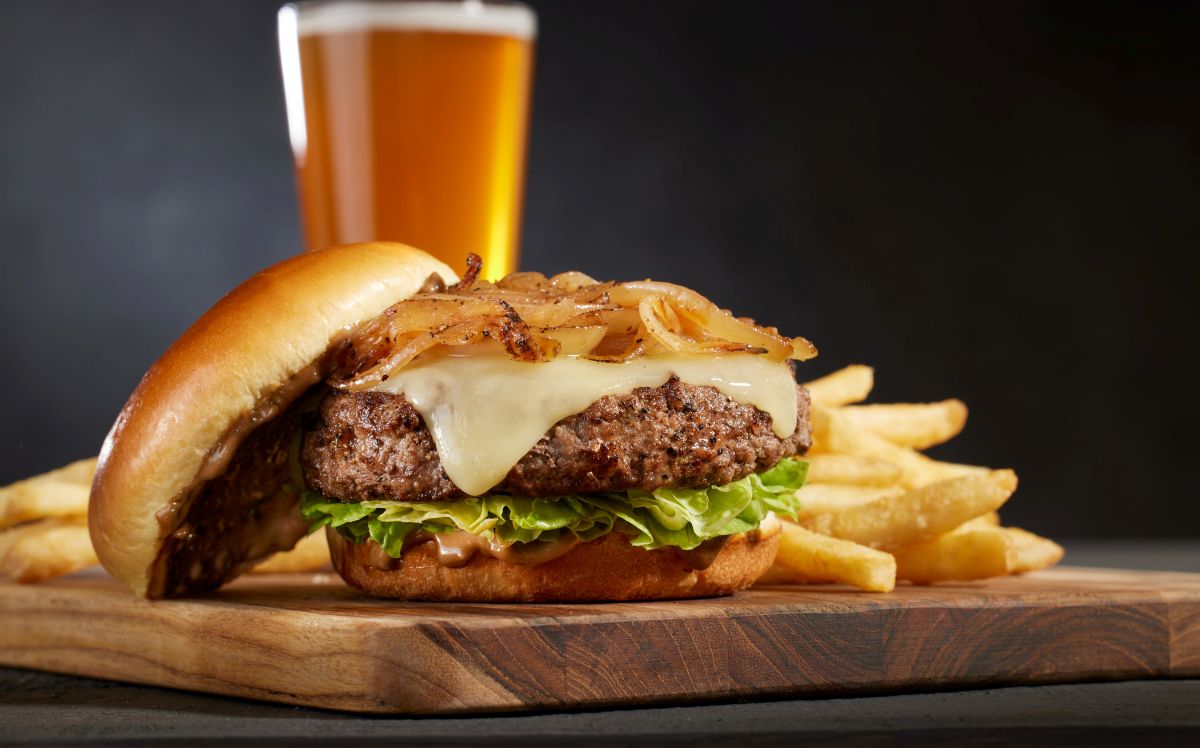 Cheeseburger with melted cheese, lettuce, and caramelized onions on a toasted bun, with fries and a beer in the background on a wooden board.