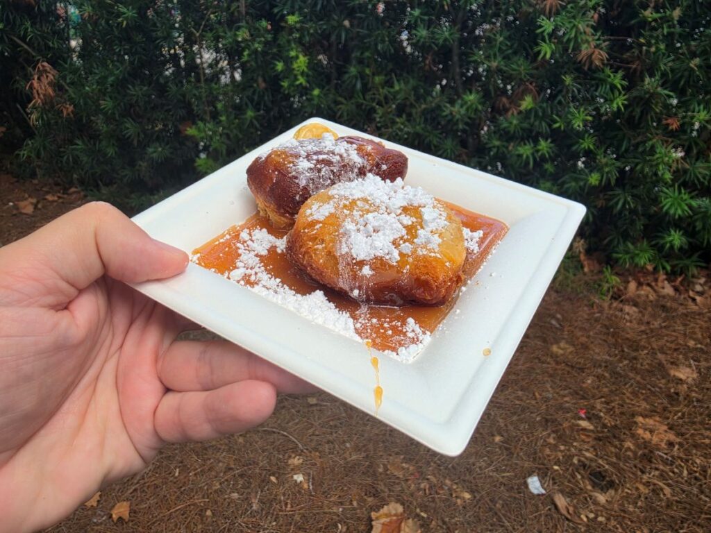 Hand holds a white square plate with two fried dough pastries dusted in powdered sugar and sitting in caramel sauce, outdoors in front of green bushes.